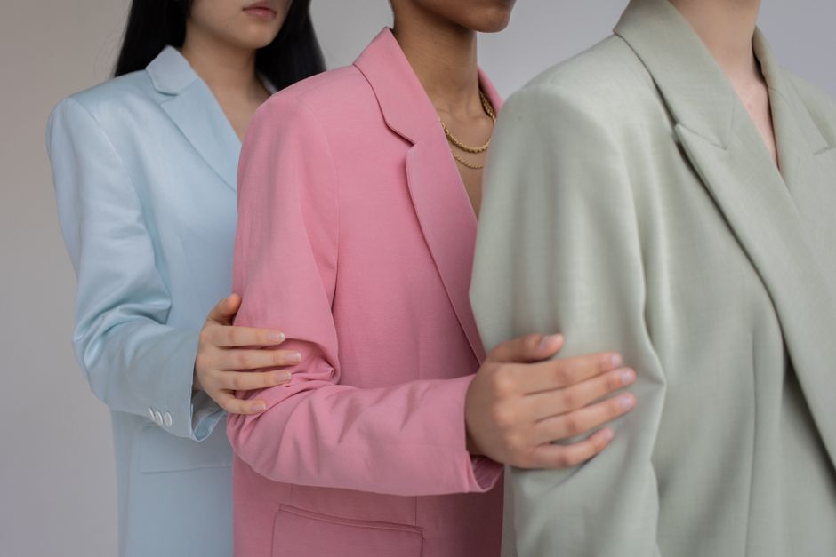 Side view of crop anonymous multiethnic female models wearing classy blazers in different pastel colors holding each others shoulders against gray background