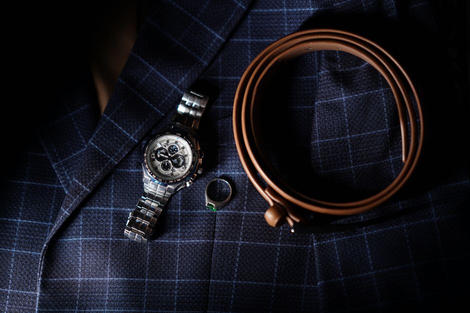 Stylish men's accessories including a wristwatch, leather belt, and silver ring on a plaid blazer.